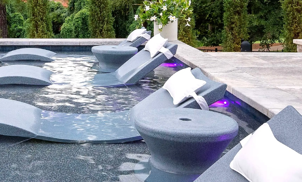 Modern in-pool chaise lounges with side tables and accent lighting designed for luxury resort and residential pool experiences.